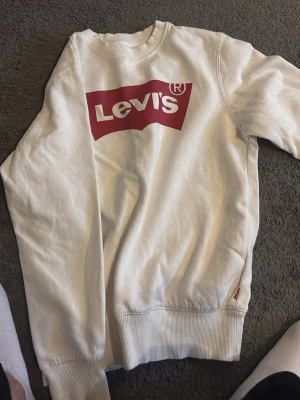 Vit sweatshirt från Levi's - Säljer en klassisk vit sweatshirt från Levi's med det ikoniska röda logotrycket på bröstet. Tröjan har rund halsringning, ribbade muddar och är tillverkad i mjukt bomullsmaterial. Perfekt för en avslappnad och stilren look.