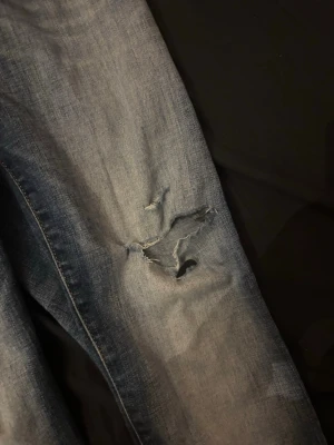 Slim fit jeans - Säljer ett par blå slim fit jeans i modellen Glenn med slitning och hål på ena benet. Jeansen har klassisk femficksdesign och är tillverkade i denim med en snygg tvättad look. Perfekta för dig som gillar en avslappnad och trendig stil.