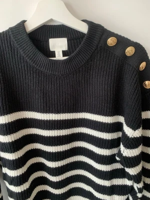 Randig stickad tröja från H&M - Snygg svart stickad tröja från H&M med vita horisontella ränder och guldfärgade knappar på ena axeln. Tröjan har rund halsringning och är ribbstickad för extra komfort. Perfekt för dig som gillar klassisk men trendig stil. Tog aldrig på mig den!