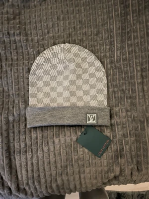 Grå Louis Vuitton mössa med rutmönster - Snygg grå mössa från Louis Vuitton med klassiskt rutmönster och vikt kant. LV-logga framtill på kanten och diskret etikett på insidan. Perfekt accessoar för dig som vill ha en exklusiv och stilren look.