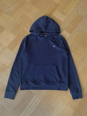 Mörkblå hoodie från GANT - Mörkblå hoodie från GANT med klassisk huva och magficka. Liten broderad logga på bröstet och ribbade muddar vid ärmslut och nederkant. Perfekt för en avslappnad och stilren look.