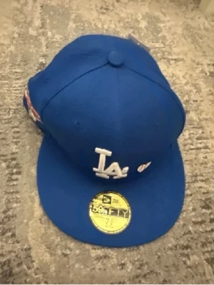 Blå LA Dodgers keps New Era 59FIFTY - Snygg blå keps från New Era 59FIFTY med LA Dodgers-logga framtill och broderad text 'LOVE & HUSTLE = GAME' på sidan. Klassisk rak skärm och MLB-logga bak. Tillverkad i slitstark bomull, perfekt för dig som gillar sportig stil.