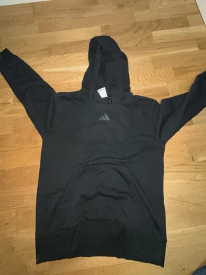 Svart hoodie från Adidas med tryck - Säljer en svart hoodie från Adidas med klassisk huva och magficka. Framtill finns Adidas-loggan i grått och på ryggen ett stort gult och svart tryck. Perfekt för dig som gillar streetwear och vill ha en skön och stilren hoodie.