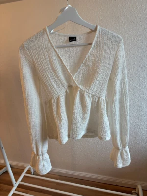 Vit omlottblus med struktur - Säljer en vit omlottblus med lång ärm och volang vid ärmslut och midja. Blusen har en snygg struktur i tyget och djup v-ringning. Perfekt för dig som gillar en trendig stil. Materialet känns mjukt och luftigt. Använd ca 3-4 gånger och är som ny. Nypris 300 kr, mitt pris 149 kr. 