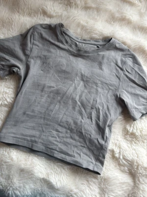 Grå basic t-shirt - Enkel och stilren grå t-shirt i bomull med rund halsringning och korta ärmar. Denna T-shirt är ganska kort i midjan. Använd några gånger men märket har ändå lyckats ”fada” bort🥰