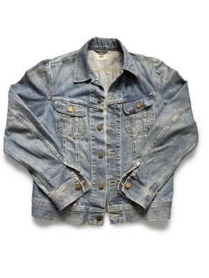 Klassisk blå jeansjacka från Lee - Snygg blå jeansjacka från Lee med klassisk passform. Jackan har två bröstfickor med lock och knappar, samt metallknappar framtill och vid ärmslut. Tidlös modell i slitstarkt denimtyg som passar perfekt till alla dina outfits. SIZE L MEN PASSAR M