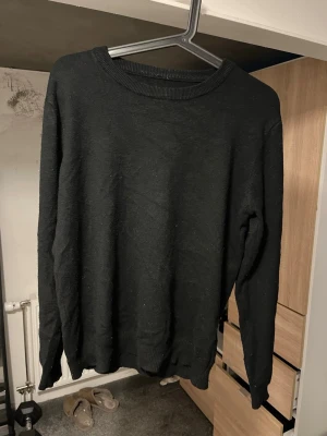 Svart stickad tröja unisex - En enkel svart stickad tröja med rund halsringning och långa ärmar. Tröjan har ribbade muddar vid hals, ärmslut och nederkant. Perfekt basplagg för lager på lager och passar till det mesta.