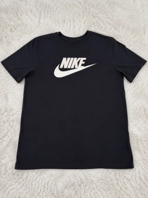 Svart Nike t-shirt med logga - Snygg svart t-shirt från Nike med klassisk vit logga på bröstet. T-shirten har rund hals och korta ärmar. Tillverkad i mjuk bomull som känns skön mot huden. Perfekt för dig som gillar sportig och enkel stil.