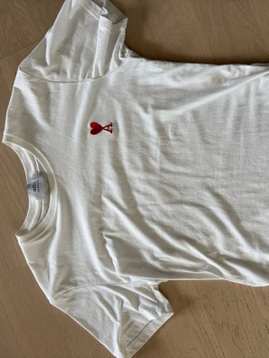 Vit t-shirt från Ami med rött hjärta - Säljer en stilren vit t-shirt från Ami Alexandre Mattiussi med ett broderat rött hjärta och bokstaven A på bröstet. Klassisk rund hals och korta ärmar. Tillverkad i mjuk bomull för en skön känsla. Tröjan är i mycket bra skick då den har få tecken på användning och inga defekter.