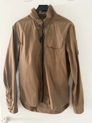 Brun overshirt med dragkedja - Snygg brun overshirt med hög krage och hel dragkedja framtill. Skjortan har en bröstficka med lock, långa ärmar med knappslå och en avslappnad passform. Tillverkad i ett slitstarkt bomullstyg som passar perfekt till lager-på-lager-stil.