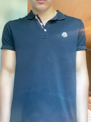 Svart Moncler pikétröja - Snygg mörkblå pikétröja från Moncler med klassisk krage och knappar framtill. Tröjan har Moncler-logga på bröstet och är tillverkad i bomull. Perfekt för en stilren och avslappnad look. Väldigt fint skick, ljuset får tröjan och se fläckig ut men tröjan är i fint skick. Äkta. Pris kan diskuteras:)
