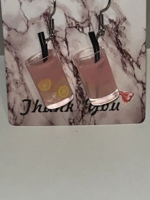 Örhängen med rosa drink och citron - Unika örhängen formade som små glas med rosa dryck, svarta sugrör och citronskivor inuti. Perfekta för dig som gillar roliga och kreativa accessoarer.Örhängens krokarna är i rostfritt stål 