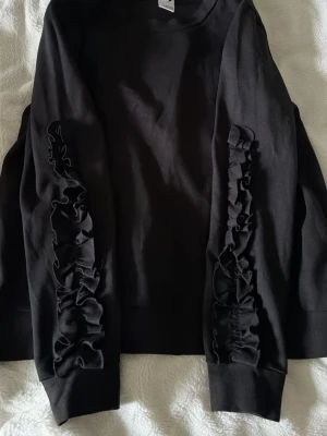 Svart sweatshirt med volangärmar - Säljer en svart sweatshirt i mjukt bomullsmaterial med volangdetaljer längs båda ärmarna. Tröjan har rund halsringning och ribbade muddar vid ärmslut och nederkant. Enkel men ändå unik tack vare volangerna på ärmarna.