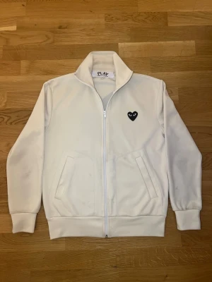 Commes des Garçons Play - Säljer en vit hoodie med dragkedja och två fickor framtill. På bröstet finns ett svart hjärta med ögon och en större version av samma hjärta på ryggen. Hoodien har ribbade muddar och hög krage. Perfekt för dig som gillar stilrena plagg med unik detalj.