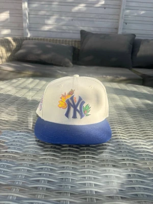 Vit och blå NY keps med brodyr - Säljer en helt ny vit keps med blå skärm och New York Yankees-logga framtill. Kepsen har broderade blommor i gult och grönt samt ett färgglatt '70th World Series'-märke på sidan. Snygg streetwear-vibe och passar perfekt till en avslappnad stil.