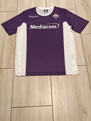 Lila och vit Kappa Fiorentina tröja - Snygg lila och vit fotbollströja från Kappa med Fiorentina-logga på bröstet och Mediacom som sponsortryck. Tröjan har korta ärmar och sportig passform, perfekt för träning eller match. Tillverkad i lätt och snabbtorkande material.