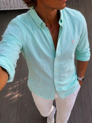 Miny Ralph Lauren skjorta med knappkrage - Snygg grön / mint Ralph Lauren skjorta med klassisk knappkrage och långa ärmar. Skjortan har knappar framtill och en liten broderad logga på bröstet. Tillverkad i bomull för en skön och luftig känsla. Perfekt för en clean och fräsch stil.