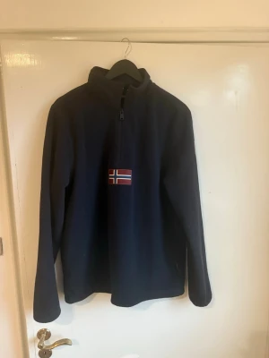 Napajiri mörkblå fleece tröja  - Mysig mörkblå fleece tröja med halv dragkedja och norsk flagga broderad på bröstet. Tröjan har hög krage och lång ärm, perfekt för kyliga dagar. Liten patch på ärmen för extra detalj.