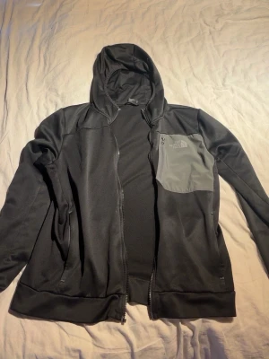 Svart The North Face hoodie - Säljer en svart hoodie från The North Face med dragkedja och huva. Den har en grå bröstficka med dragkedja och logga på vänster sida. Materialet är mjukt och stretchigt, perfekt för chill eller träning. Dragkedjan är lite seg och kan fastna ibland men utöver det så är den som ny