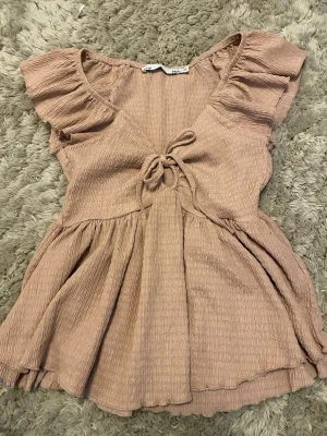 Beige omlottopp med volangärm - Supersöt beige omlottopp med volangärmar och knyt framtill. Toppen har en lätt rynkad struktur och är tillverkad i ett mjukt material som känns skönt mot huden. Perfekt för dig som gillar romantiska detaljer och en feminin look.