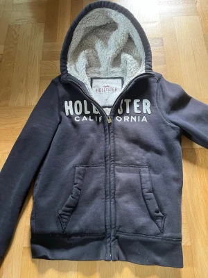 Svart hoodie från Hollister med foder 2000s - Mysig svart hoodie från Hollister med dragkedja och vit teddyfoder i hela hoodie. Stor logga framtill med texten 'Hollister California'. Två fickor fram och ribbade muddar. Perfekt för kyliga dagar. Vintage