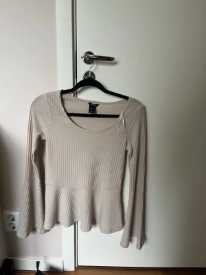 Beige ribbad topp med utsvängda ärmar - Säljer en beige ribbad topp med utsvängda långa ärmar och peplum nedtill. Toppen har rund halsringning och är tillverkad i ett mjukt, stretchigt material som sitter snyggt på kroppen. Perfekt för dig som gillar en stilren men ändå unik look.