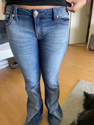 Blå bootcut jeans True Religion - Säljer ett par blå bootcut jeans från True Religion med röda kontrastsömmar och klassiska fickor bak. Jeansen har låg midja och är tillverkade i denim med lätt tvättad look. Perfekta för dig som gillar en avslappnad men trendig stil.