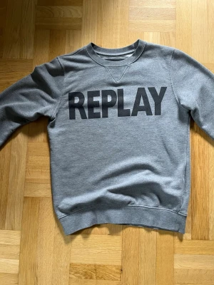 Grå sweatshirt från Replay - Snygg grå sweatshirt från Replay med stor svart logga över bröstet. Klassisk rund halsringning och ribbade muddar vid ärmslut och nederkant. Perfekt för en avslappnad och trendig look. Använd max 2 gånger. Den har ingen storlekstag men den är typ S till liten M. 