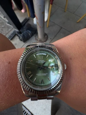 R klocka med grön urtavla - Snygg R klocka med grön urtavla och silverfärgat armband i metall. Klockan har romerska siffror, datumvisning och dagvisning. Klassisk räfflad bezel och robust länkarmband ger en exklusiv känsla.