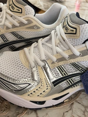 Vita Asics sneakers med mesh och silver - Snygga vita Asics sneakers med mesh-överdel, detaljer i silver och beige samt svarta inslag. Skorna har en sportig look med tjock sula och tydliga loggor på sidan och plösen. Perfekta för dig som gillar streetwear och komfort. Klassisk snörning och ventilerande material.