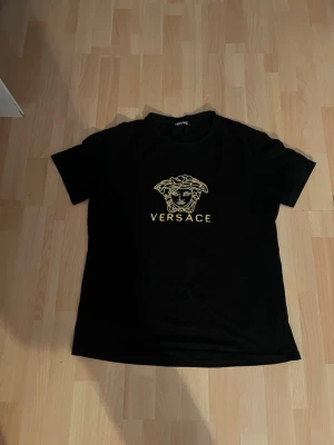 Svart t-shirt med guldtryck - Snygg svart t-shirt med ikoniskt Medusa-huvud och logga i guld på bröstet. Klassisk rund halsringning och korta ärmar. Perfekt för dig som vill ha en stilren och exklusiv look.