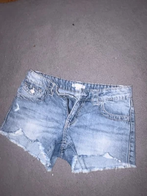 Blå jeansshorts med rå kant - Säljer ett par blå jeansshorts med fransig, rå kant och slitna detaljer. Shortsen har klassisk femficksdesign, bälteshällor och knappgylf. Perfekta för sommaren och ger en avslappnad vibe.