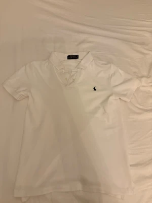 Vit pikétröja från Polo Ralph Lauren - Klassisk vit pikétröja från Polo Ralph Lauren med korta ärmar och krage. Tröjan har knappar vid halsen och den ikoniska broderade loggan på bröstet. Perfekt till jeans eller shorts för en clean och stilren look.