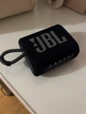 JBL svart bärbar högtalare - Snygg och kompakt svart bärbar högtalare från JBL. Högtalaren har en robust tygklädd yta, tydliga knappar på ovansidan och en praktisk bärrem. Perfekt för att ta med sig musiken överallt och enkel att koppla till mobilen via Bluetooth.