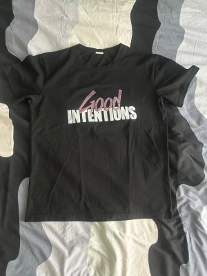 Svart t-shirt med tryck Good Intentions - Säljer en svart t-shirt med färgglatt tryck 'Good Intentions' på framsidan och ett stort lila V med vita duvor på baksidan. T-shirten har rund hals och korta ärmar. Perfekt för dig som gillar streetwear och grafiska prints.