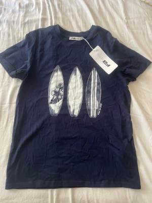 Mörkblå t-shirt med surfbrädor - Snygg mörkblå t-shirt från Sister med tryck av tre surfbrädor på bröstet, varav en har blommönster. T-shirten har rund hals och korta ärmar. Perfekt för en avslappnad och cool stil.