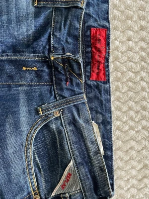 Replay Jeans - Snygga blå jeans från Replay med klassisk femficksmodell och coola röda detaljer på etiketten och fickan. Jeansen har gul kontrastsöm och en lätt tvättad look som ger en avslappnad vibe. Perfekta för dig som gillar streetwear och vill ha något stilrent.