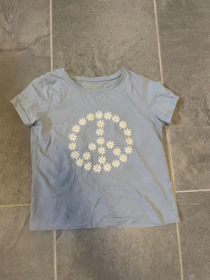 Ljusblå t-shirt med peace-märke - Ljusblå t-shirt från Primark med korta ärmar och ett stort peace-märke på bröstet, gjort av vita blommor. Perfekt för en avslappnad och cool stil. Tillverkad i mjuk bomull för skön känsla hela dagen.