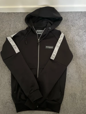 Svart hoodie från McKenzie med dragkedja - Snygg svart hoodie från McKenzie med dragkedja framtill och huva med dragsko. Gråa detaljer med McKenzie-tryck längs ärmarna ger en sportig vibe. Tillverkad i mjukt material och har fickor med dragkedja på sidorna. Perfekt för en chill och avslappnad stil.