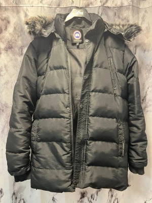 Svart dunjacka med pälskantad huva - Snygg svart dunjacka från Canada Goose med pälskantad huva och quiltad design. Jackan har dragkedja och knappar framtill, flera fickor och ribbade muddar vid ärmsluten. Perfekt för kalla dagar när du vill hålla dig varm och stilren.