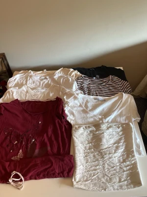6-pack t-shirts i olika färger och mönster - Säljer ett paket med sex t-shirts i olika färger och stilar. Innehåller flera vita t-shirts, en vinröd med fjärilstryck, en svart och en vit/svart-randig. Alla har rund hals och korta ärmar, perfekta för en enkel och avslappnad look.
