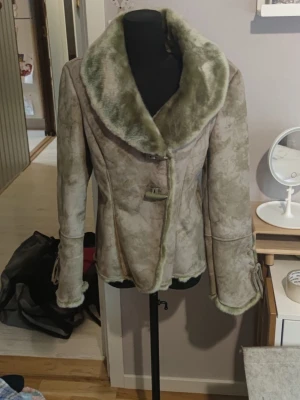 Olivgrön kappa med fuskpäls - Säljer en olivgrön kappa med bred krage och detaljer i grön fuskpäls. Kappan har markerad midja, dekorativa spännen och långa ärmar med pälsdetaljer vid ärmslut. Perfekt för dig som vill ha en unik och trendig look.