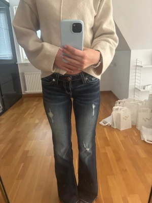 Mörkblå bootcut jeans med slitningar - Säljer ett par mörkblå bootcut jeans med vita kontrastsömmar och slitna detaljer på benen. Jeansen har låg midja, fem fickor och röda detaljer vid fickorna. Perfekta för en avslappnad och trendig look. Storlek 152, är 160 lång och passar mig toppenbra! 