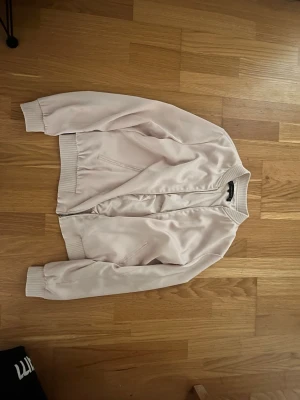Benvit satin bomberjacka - Snygg benvit bomberjacka i satin med ribbade muddar vid ärmslut, krage och nederkant. Jackan har dragkedja framtill och två sidofickor. Perfekt för dig som vill ha en stilren och trendig look.