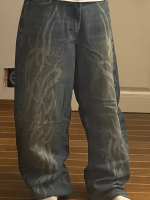 Baggy jeans med tribal-mönster - Säljer ett par riktigt baggy jeans från FBsister i tvättad blå denim med stora, ljusa tribal-mönster längs båda benen. Jeansen har fem fickor, hög midja och bred passform för en skön streetwear-look. Perfekta för dig som vill sticka ut!