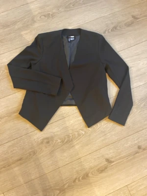 Svart kavaj från H&M Divided - Stilren svart kavaj från H&M Divided i storlek 34. Kavajen har en öppen front utan knappar, lång ärm och är helfodrad. Passformen är rak och enkel, vilket gör den lätt att matcha med olika outfits. Materialet känns mjukt och följsamt.