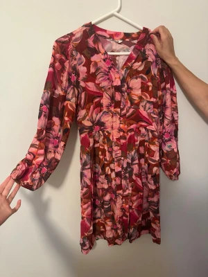 Blommig klänning i rosa och rött - Säljer en färgstark klänning med blommigt mönster i rosa, rött och brunt. Klänningen har v-ringning, knappar framtill och långa ärmar med resår vid ärmslut. Perfekt för dig som gillar statement-plagg och vill sticka ut. Storlek S men passar även M