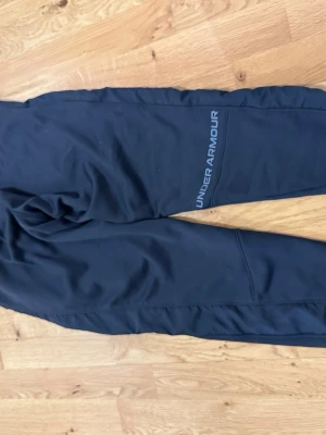 Svarta mjukisbyxor från Under Armour - Snygga svarta mjukisbyxor från Under Armour med logga längs benet. Tillverkade i ett mjukt och stretchigt material som passar perfekt för chill eller träning. Byxorna har en avslappnad passform och resår i midjan.