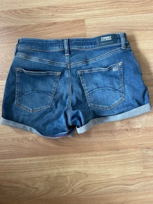 Blå jeansshorts från Tommy Jeans - Snygga blå jeansshorts från Tommy Jeans med klassisk femficksdesign och uppvikta benslut. Shortsen har en mörkblå tvätt, bakfickor och diskret logga på ena fickan. Perfekta för varma dagar och enkel att matcha med olika stilar.