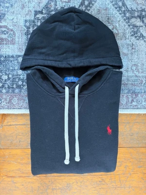 Svart Polo Ralph Lauren Hoodie  - Hej! 👋 Säljer en klassisk svart Polo Ralph Lauren hoodie i storlek M med röd pony-logga. 🐎✨ Något bredare/rymligare modell! Fint skick överlag men är något solblekt, bland annat på magfickan (se bilder). Inga hål eller skador. Garanterat 100% äkta med alla originaltvättlappar kvar (ingen QR-kod på denna årsmodell). Ett perfekt och riktigt snyggt basplagg! Fraktar snabbt! 💯✅🚛📦💬 